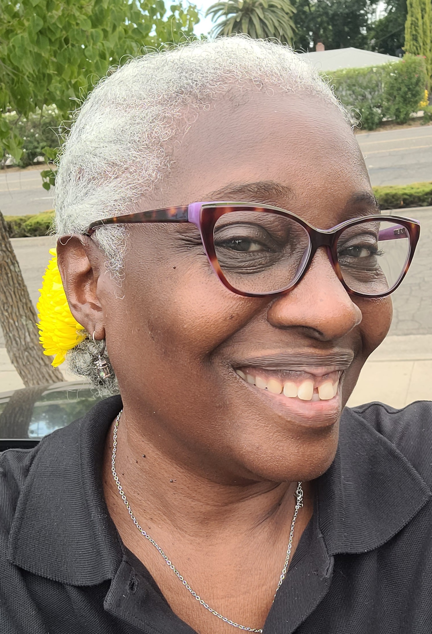 Tanya pic A woman with short gray hair and glasses smiles outdoors, wearing a black shirt and a yellow flower behind her ear.