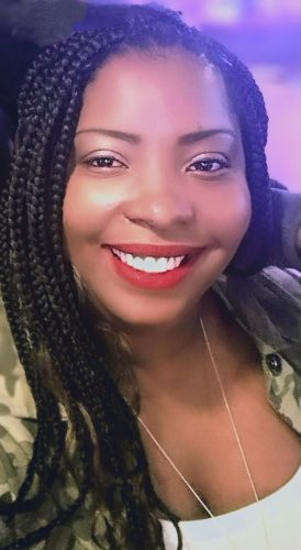 CHMACY Board Member A woman with braided hair and red lipstick smiles at the camera, wearing a camouflage jacket and a white top.