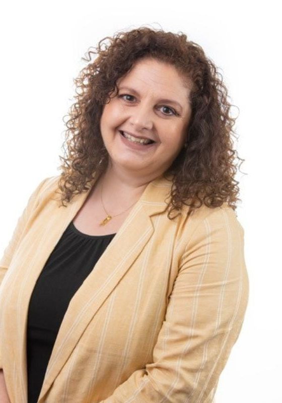 A person with curly hair smiles while wearing a yellow blazer over a black top.