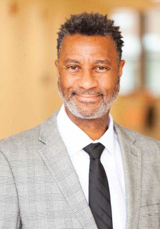 A man with short curly hair and a beard is wearing a gray checked suit jacket, white shirt, and black tie. He is standing against a blurred indoor background.